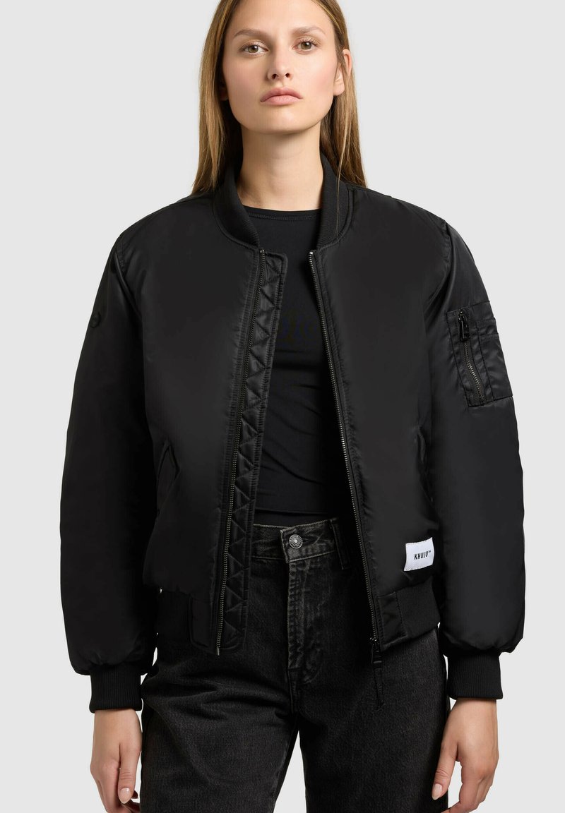 Giacca bomber nera con chiusura a zip, polsini a costine e patch con logo cucito. Il materiale è lucido e imbottito per garantire calore. Presenta una tasca laterale.