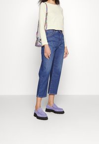 Gestreiftes gelbes und weißes Langarmoberteil, blaue hochtaillierte Cropped-Jeans, lila Loafers mit dicken schwarzen Sohlen und eine gemusterte Schultertasche.