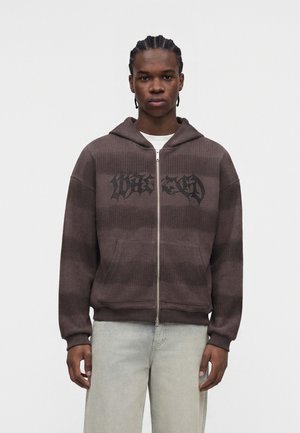 Jeune homme aux cheveux tressés porte un sweat à capuche marron zippé avec un texte noir et un pantalon gris clair, debout devant un fond uni.