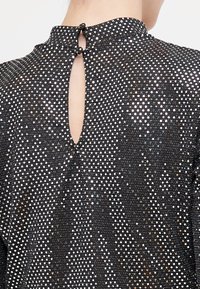 Haut noir avec un col haut, doté d'une fermeture au dos en forme de trou de clé. Le tissu présente un motif texturé avec des points en relief brillants sur l'ensemble.