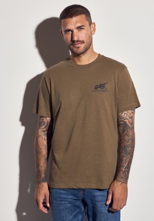 Mand med kort hår og fuld arm tatovering iført brun t-shirt og blå jeans, stående mod en hvid væg og kaster en skygge.