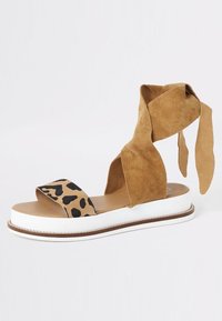 Ruskeat mokkasaappaat, joissa on leopardi-kuvioinen hihna, avoin varvas, paksu valkoinen pohja ja pitkät mokkarenkaat, jotka kietoutuvat nilkan ympäri.