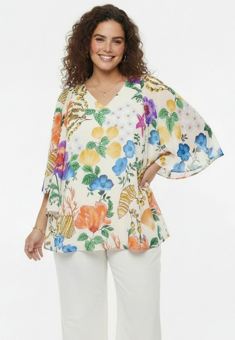 Glimlachende vrouw met krullend haar, gekleed in een losse, bloemrijke blouse met wijde mouwen en een witte broek, tegen een effen achtergrond.