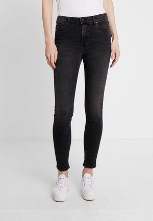 Femme portant un jean noir slim et des baskets blanches, debout devant un fond gris clair uni.