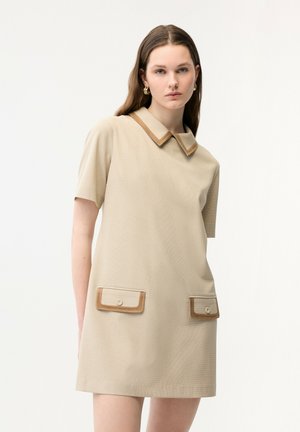 Frau trägt ein beige kurzärmeliges Kleid mit kontrastierendem Kragen und zwei Vordertaschen, steht vor einem schlichten weißen Hintergrund.