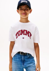Hvit bomull t-skjorte med en rød brodert "TOMMY" logo, brukt sammen med blå denimjeans og en marineblå cap med hvit tekst.