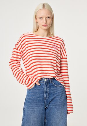 Blonde Frau, die ein rot-weiß gestreiftes Langarmshirt und hoch taillierte blaue Jeans trägt, mit einer Hand in der Tasche.