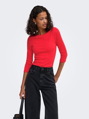 Vrouw met krullend haar die een rood nauwsluitend topje en zwarte high-waisted jeans draagt, met in één hand een zwarte handtas tegen een effen achtergrond.