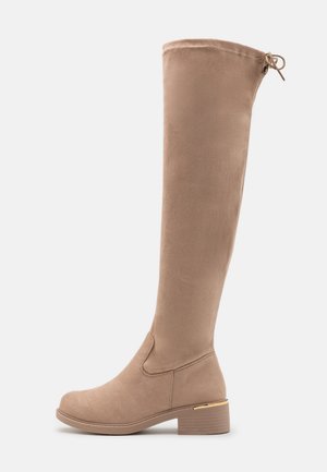 Beige suède kniehoog laars met lage blokhak en strikdetail aan de achterkant, zijaanzicht op witte achtergrond.