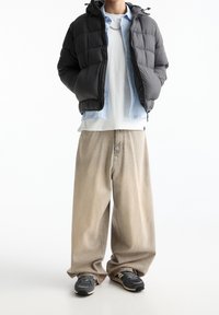 Gepolsterte schwarze Jacke mit Kapuze, blaues Hemd zum Knöpfen, weißes T-Shirt und locker sitzende beige Hose, kombiniert mit schwarzen Turnschuhen.