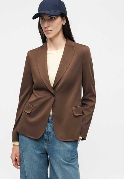 Γυναίκα που φοράει καφέ σακάκι πάνω από ανοιχτό κίτρινο τοπ, μπλε τζιν και μπλε σκούφο μπέιζμπολ, στέκεται με το ένα χέρι στην τσέπη.