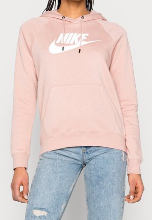 Personne portant un sweat à capuche Nike rose clair avec un logo blanc et un jean déchiré bleu clair, debout devant un fond uni.
