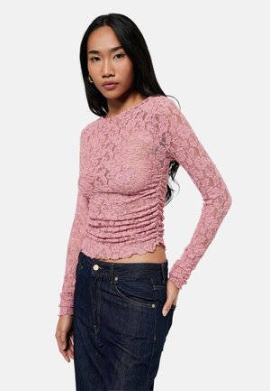 Mujer con cabello largo y negro lleva una blusa rosa de encaje de manga larga y vaqueros azul oscuro, posando con una mano en el bolsillo contra un fondo blanco.