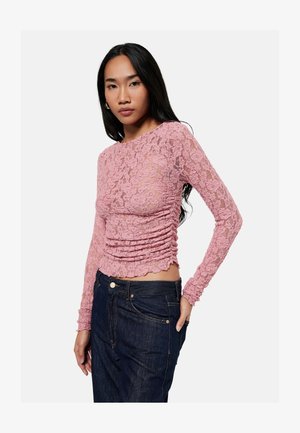 Mujer con cabello largo y negro lleva una blusa rosa de encaje de manga larga y vaqueros azul oscuro, posando con una mano en el bolsillo contra un fondo blanco.
