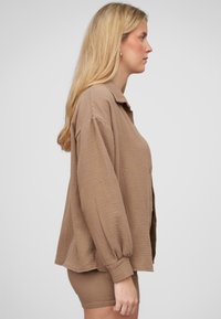 Camicia di un marrone chiaro, con texture, colletto e maniche lunghe a sbuffo, abbinata a un shorts coordinato.