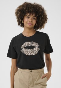 Zwarte katoenen t-shirt met een lippenafbeelding in luipaardprint. Korte mouwen, ronde hals, ontspannen pasvorm, gladde textuur. G gedragen met beige broek.