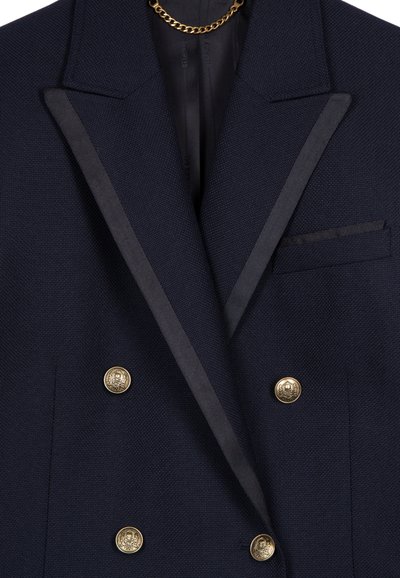Blazer doppiopetto blu navy con bottoni dorati, con rever in satin e interno foderato. Presenta una tasca sul petto e tessuto testurizzato.