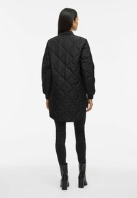 Γυναίκα ντυμένη με ένα μακρύ μαύρο επενδυμένο μπουφάν, μαύρες κολάν και μαύρες γόβες με ύψος στον αστράγαλο, που στέκεται με πλάτη σε έναν λευκό φόντο.