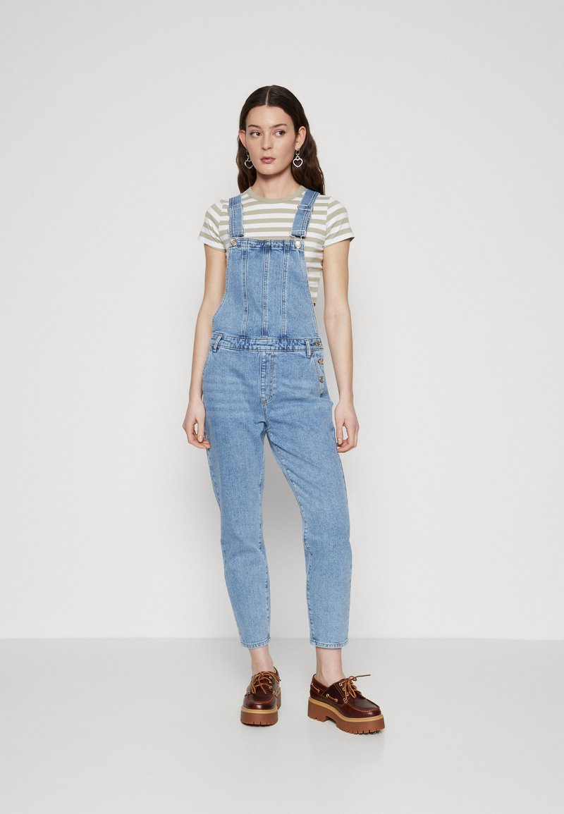 Pantalones de mezclilla azul claro con tirantes ajustables y un ajuste entallado, combinados con una blusa de rayas de manga corta y zapatos de plataforma marrones.