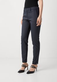 Marinblå skräddarsydda byxor i en slät tyg, slim fit och subtila veck; ihop med svarta spetsskor med spetsiga tår som har guldiga cirkulära detaljer.