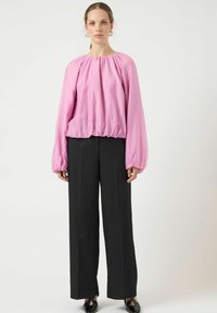 Lyserød bluse med ballonærmer og rynket halsudskæring, kombineret med højtaljede sorte bukser med lige ben og slidser.