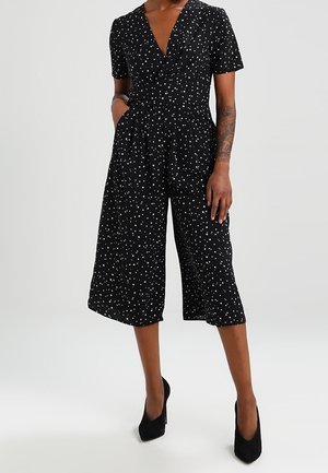 Combinaison noire à manches courtes avec des pois blancs, col en V, jambes larges coupées au milieu du mollet, portée avec des talons hauts noirs.