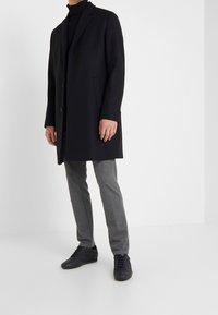 Zwarte wolblend overjas met een geknipte kraag, enkele knoopsluiting, zijzakken, gedragen over een zwarte coltrui en grijze trousers. Zwarte sneakers.