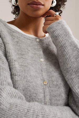 Isik, kes kannab pehmet halli kootud kardigani pärlknappidega, hõbedast rõngakujulisi kõrvarõngaid ja hõbedast sõrmust, käsi puhkab lõua lähedal.