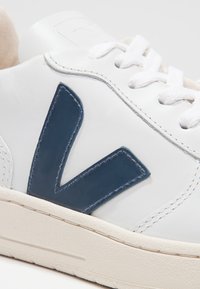 Gros plan sur une basket blanche avec un logo en forme de V bleu foncé sur le côté et des lacets blancs sur une semelle claire.