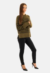Maglione a righe verde oliva realizzato in tessuto morbido, con maniche lunghe e spacchi laterali, abbinato a pantaloni neri aderenti e tacchi alti neri.