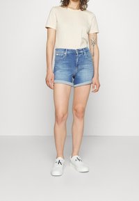 Shorts en denim bleu clair avec desourlets roulés, associés à une chemise à manches courtes crème et des baskets blanches. Tenue simple et décontractée.
