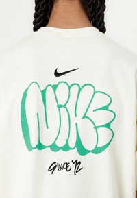 Balta kokvilnas džemperis ar lielu zaļu un melnu grafiti stila "NIKE" grafiku uz muguras, kurā rakstīts "SINCE '72" melnā krāsā.