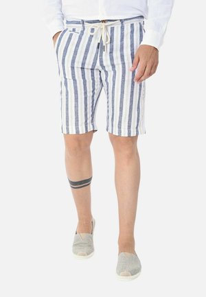 Uomo che indossa pantaloncini bianchi e blu a righe verticali con coulisse, camicia bianca, scarpe slip-on grigie e un tatuaggio alla caviglia sulla gamba sinistra.