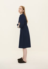 Dunkelblaues Kleid mit langen Ärmeln, tailliertem Bund und Midi-Länge. Getragen mit dunkelbraunen Clogs. Einfacher Hintergrund, Model steht seitlich.