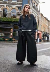 Sötétszürke, bő méretű kötött pulóver gallérral, laza fekete, széles szárú nadrággal és fekete lapos cipővel, szabadtéren, városi környezetben.