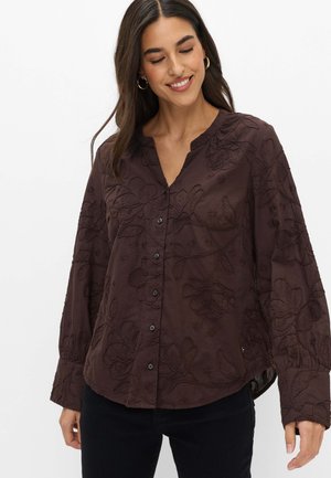 Vrouw draagt een donkerbruine blouse met lange mouwen en bloemenborduursel, gecombineerd met een zwarte broek, glimlachend en naar beneden kijkend.