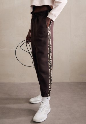 Personne portant un pantalon de survêtement Adidas marron foncé avec des bandes latérales à imprimé animal, des chaussettes blanches Adidas et des baskets, tenant une corde à sauter.
