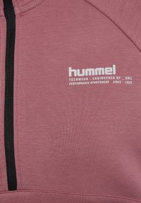 Mittelschwerer, rosa Techwear-Hoodie mit strukturierter Oberfläche, ausgestattet mit einem schwarzen Reißverschluss und einem auffälligen weißen Logo auf der linken Seite.