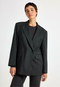 Sort blazer med pinstriber, strukturerede skuldre, dobbelt brystlås og to frontlommer. Båret over en sort top med en glat tekstur.