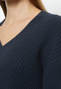 Pull côtelé bleu marine avec un col en V. Le tissu a une finition douce et texturée, avec des côtes verticales et une coupe décontractée.