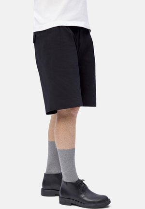 Hombre vestido con pantalones cortos negros, calcetines grises hasta la mitad de la pantorrilla y botas negras de cordones hasta el tobillo, de pie contra un fondo blanco liso.