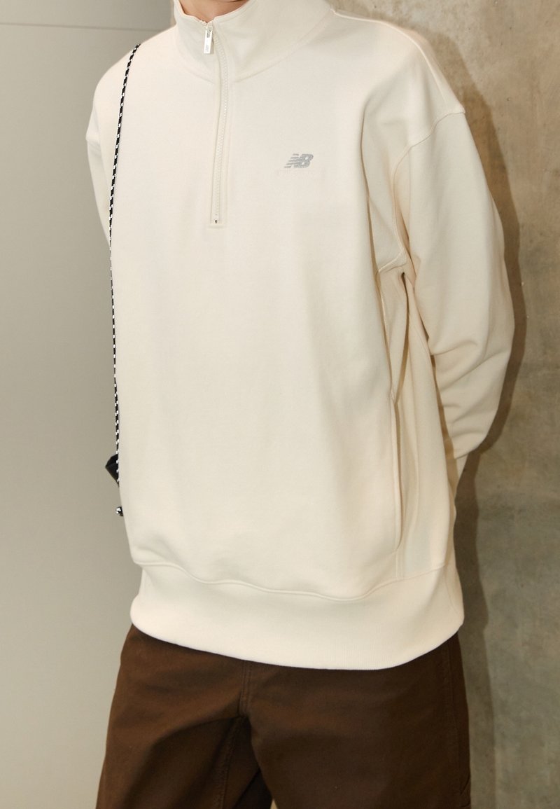 Personne portant un sweat-shirt New Balance crème avec zip quart et un pantalon marron, debout contre un mur uni, les mains dans le dos.