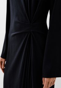 Abito nero realizzato in un tessuto liscio e elastico, con un dettaglio a torsione in vita e sottili pieghe drappeggiate lungo il lato. Maniche lunghe.