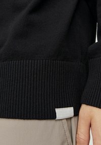 Czarny dzianinowy sweter z prążkowanym dołem. Miękka textura, posiadająca małą metkę tkaninową w dolnym rogu. Nosi się go na beżowych spodniach.
