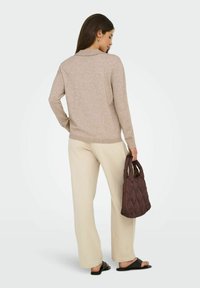 Mujer de pie, vistiendo un suéter beige y pantalones, sosteniendo un bolso acolchado marrón, con sandalias negras de llevar, sobre un fondo blanco.