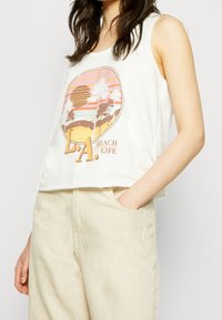 Crop top bianco senza maniche con una grafica di un tramonto con palme e il testo "L.A. BEACH LIFE"; abbinato a pantaloni beige a vita alta.