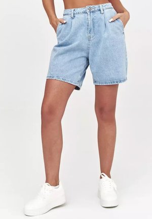 Kobieta w jasnoniebieskich krótkich spodenkach dżinsowych z wysokim stanem i przednimi kieszeniami oraz białych trampkach, stoi z rękami w kieszeniach.