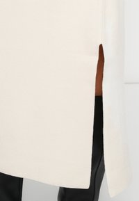 Pull en tricot couleur crème avec fentes latérales, présentant une texture lisse et un ourlet droit. Pantalons noirs visibles sous le pull.