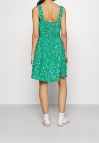 Grünes Blumenmusterkleid mit tailliertem Oberteil und ausgestellt Rock. Verfügt über breite Träger und ein quadratisches Rückendesign. Getragen mit weißen Sneakern.