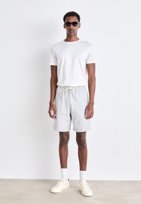 White t-shirt, gray shorts with drawstring, and beige sneakers. Model wears black sunglasses, stands against a plain backdrop.
